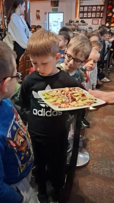 Przedszkolaki podczas wizyty w fabryce czekolady  z zaciekawieniem degustują różne smakołyki, uśmiechając się szeroko. W tle można dostrzec urządzenia i maszyny związane z produkcją czekolady, co dodaje uroku całej scenie.