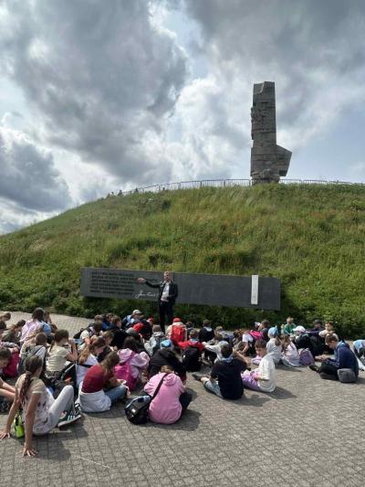 Uczniowie siedzą na ziemi przed pomnikiem na Westerplatte i słuchają przewodnika