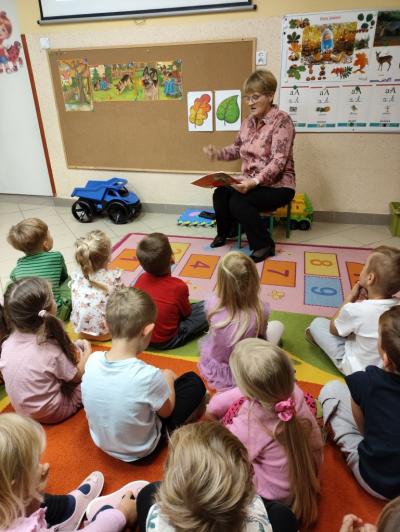Pani Bożenka czyta opowiadanie. Przed nią siedzą dzieci na dywanie. W tle tablica z ilustracjami do opowiadania.