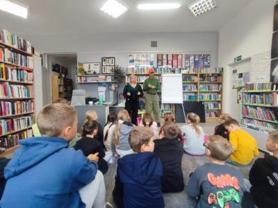 Uczniowie siedzą na dywanie, przed nimi pan z plakatem w rękach, obok stoi pani bibliotekarka.