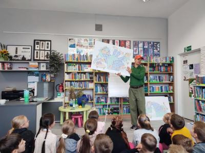 Uczniowie siedzą na dywanie, przed nimi pan z plakatem w rękach.