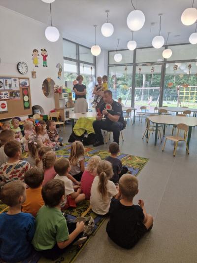 Dzieci siedzą na dywanie i słuchają Policjanta, który opowiada o bezpieczeństwie na drodze