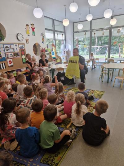 Dzieci siedzą na dywanie i słuchają Policjanta, który opowiada o bezpieczeństwie na drodze.