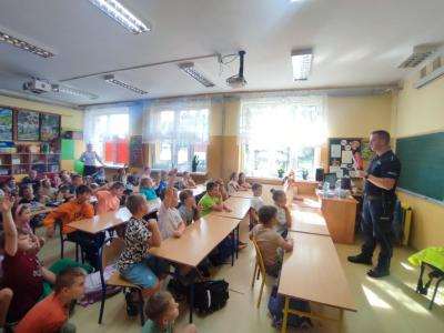 Pan policjant stoi przed ławkami, w których siedzą uczniowie.
