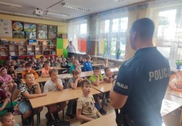 Pan policjant stoi przed ławkami, w których siedzą uczniowie. Pan policjant stoi przed ławkami, w których siedzą uczniowie.