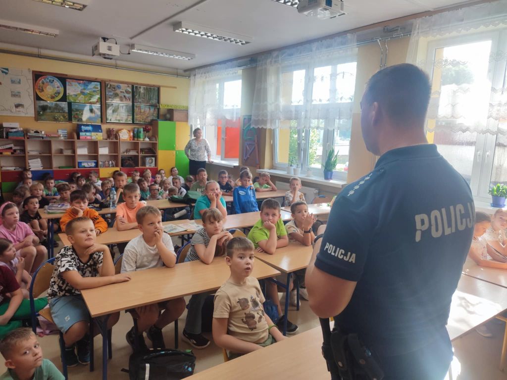 Pan policjant stoi przed ławkami, w których siedzą uczniowie.