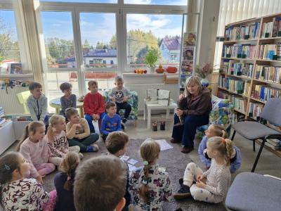 Dzieci siedzą na dywanie i słuchają opowiadania, które czyta Pani  bibliotekarka