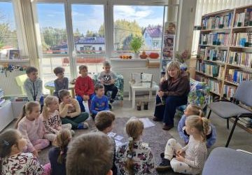 Dzieci siedzą na dywanie i słuchają opowiadania, które czyta Pani  bibliotekarka