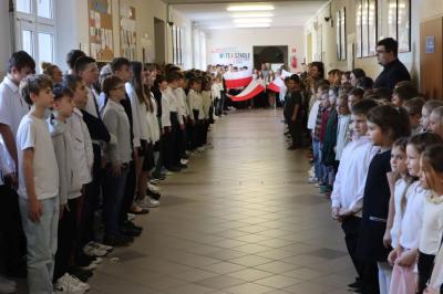 Uczniowie podczas apelu odśpiewują hymn polski. W tle flagi Polski.