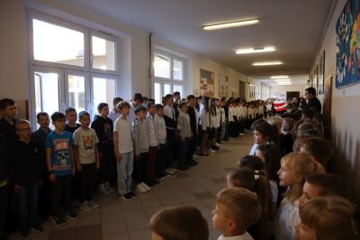 Uczniowie podczas apelu odśpiewują hymn polski. W tle flagi Polski.