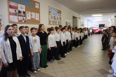 Uczniowie podczas apelu odśpiewują hymn polski. W tle flagi Polski.