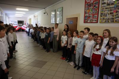 Uczniowie podczas apelu odśpiewują hymn polski. W tle flagi Polski.