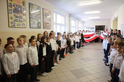 Uczniowie podczas apelu odśpiewują hymn polski. W tle flagi Polski.