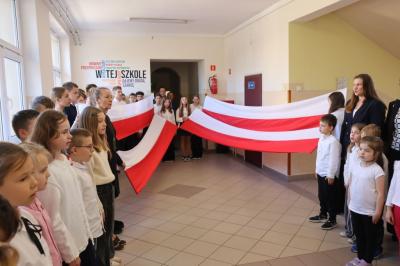 Uczniowie podczas apelu odśpiewują hymn polski. W tle flagi Polski.