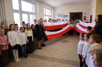 Uczniowie podczas apelu odśpiewują hymn polski. W tle flagi Polski.