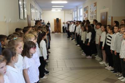 Uczniowie podczas apelu odśpiewują hymn polski. W tle flagi Polski.