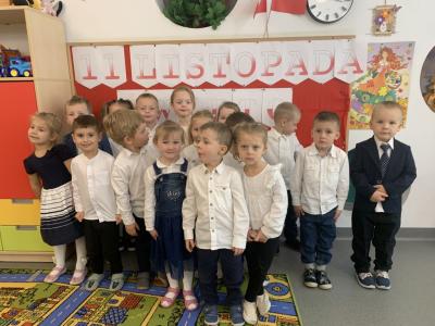 Odświętnie ubrane dzieci stoją przed tablicą z napisem 11 listopada i śpiewają Hymn państwowy.