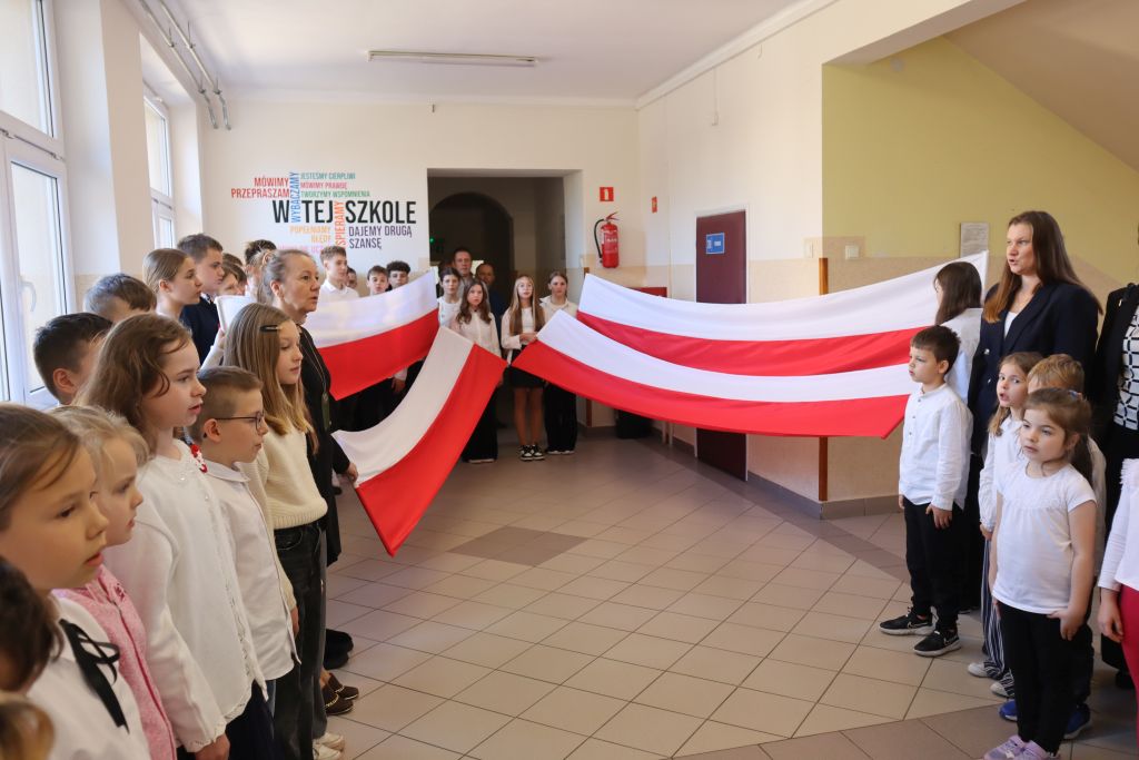 Uczniowie podczas apelu odśpiewują hymn polski. W tle flagi Polski.