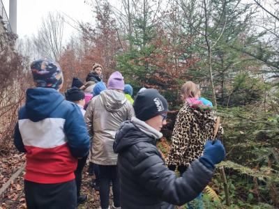 Dzieci wśród drzew, w zimowych ubraniach idą za panią przewodnik..