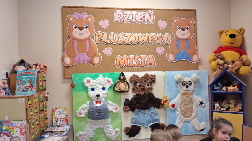 Na ścianie wisi tablica z hasłem Dzień Pluszowego Misia oraz duże ilustracje przedstawiające misie.