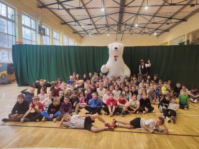 Dzieci siedzą na parkiecie. Pozują do zdjęcia. Na środku stoi duży, biały miś