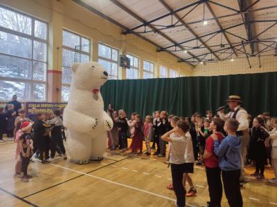 Dzieci tańczą z ogromnym, białym misiem w hali sportowej.