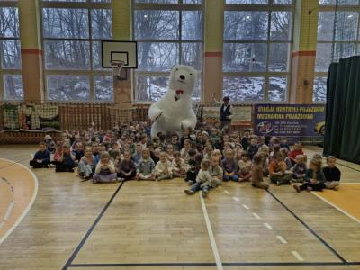 Przedszkolaki siedzą na parkiecie. Pozują do zdjęcia. Na środku stoi duży, biały miś