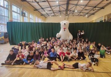 Dzieci siedzą na parkiecie. Pozują do zdjęcia. Na środku stoi duży, biały miś.