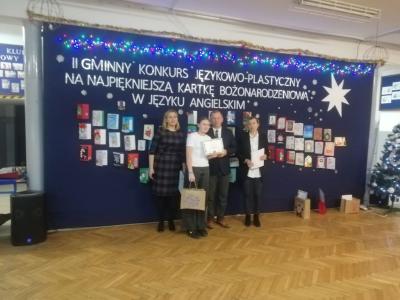 Na zdjęciu Natalia Pabis stoi przed wystawą kartkek świątecznych i trzyma w dłoni dyplom oraz nagrodę