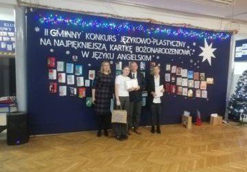 Na zdjęciu Natalia Pabis stoi przed wystawą kartkek świątecznych i trzyma w dłoni dyplom oraz nagrodę.