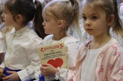 Przedszkolaki stojące przy scenie, śpiewają sto lat, w rękach trzymają upominki dla dziadków