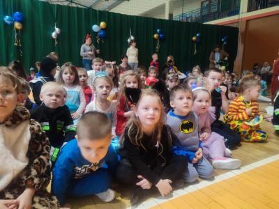 Dzieci w strojach karnawałowych siedzą na parkiecie w hali soprtowej