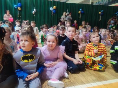 Dzieci w strojach karnawałowych siedzą na parkiecie w hali soprtowej