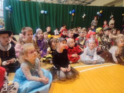 Dzieci w strojach karnawałowych siedzą na parkiecie w hali soprtowej