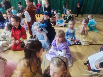 Dzieci w strojach karnawałowych siedzą na parkiecie w hali soprtowej