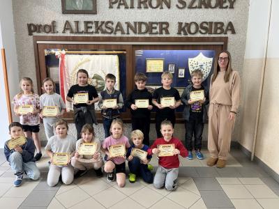 dzieci świetlicowe trzymają dyplomy, stoją w dwóch rzędach z nauczycielką obok gabloty patrona szkoły