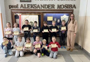 dzieci świetlicowe trzymają dyplomy, stoją w dwóch rzędach z nauczycielką obok gabloty patrona szkoły