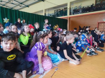 Dzieci w strojach karnawałowych siedzą na parkiecie w hali sportowej