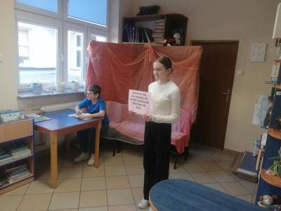 Uczennica stoi i trzyma w dłoni kartkę z życzeniem książki.