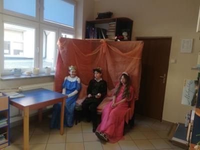 Uczniowie w strojach króla, królewny i królowej siedzą na krzesełkach.
