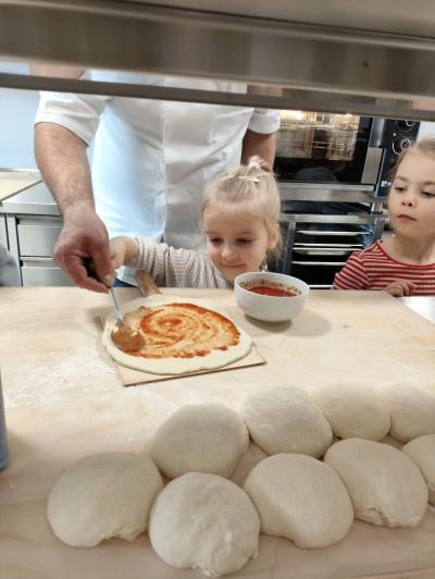 Dziewczynka z pomocą kucharza smaruje sosem ciasto do pizzzy.