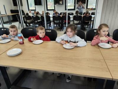 Dzieci siedzą przy stołach i degustują pizzę. W tle odbicie w lustrach.