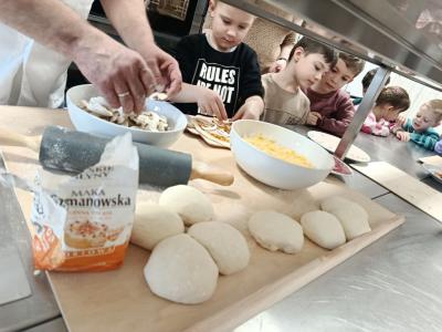 Chłopiec nakłada na pizzę składniki, a kucharz pomaga mu podając pieczarki.