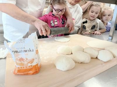 Dziewczynka wałkuje ciasto na pizzę przy pomocy kucharza, który ją instruuje