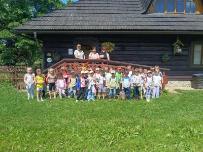 zdjęcie grupowe przedszkolaków na tle starego drewnianego domku. Dzieci stoją na trawie wraz z nauczycielkami