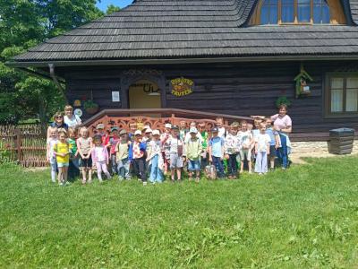 zdjęcie grupowe przedszkolaków z grup Skrzaty i Biedronki podczas wizyty w Sądeckim Batniku. Dzieci stoją na tle drewnianego domku wraz z wychowawcami