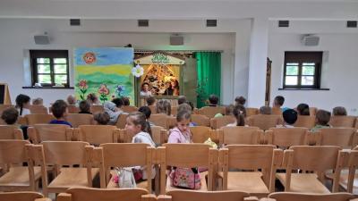 Teatrzyk kukielkowy dla dzieci w srodku w budynku. Dzieci siedzą na krzesełkach przed sceną i z zaciekawieniem obserwują co dzieje się za zieloną kotarą