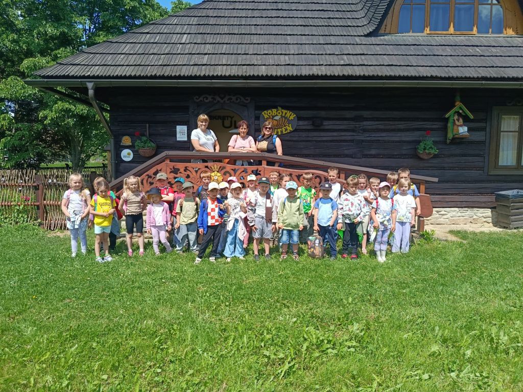 zdjęcie grupowe przedszkolaków na tle starego drewnianego domku. Dzieci stoją na trawie wraz z nauczycielkami