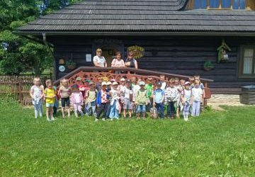 zdjęcie grupowe przedszkolaków na tle starego drewnianego domku. Dzieci stoją na trawie wraz z nauczycielkami