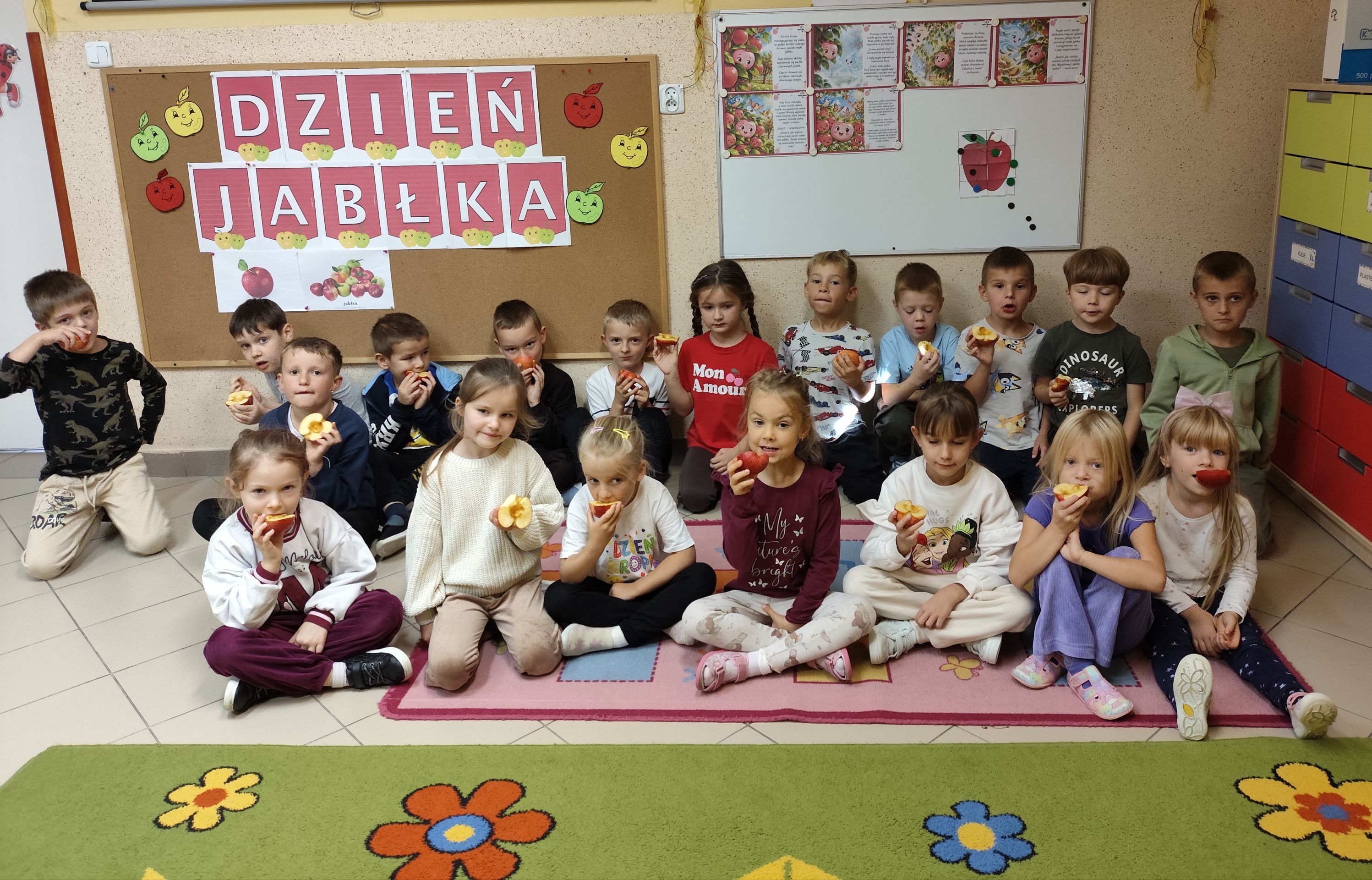 Dzieci jedzą jabłka na tle tablicy z okazji Dnia Jabłka.